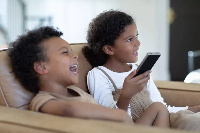 Two children laughing on a sofa while one uses a phone